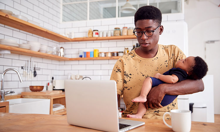 Multi-Tasking Father Holds Sleeping Baby Son And Works On Laptop Computer In Kitchen