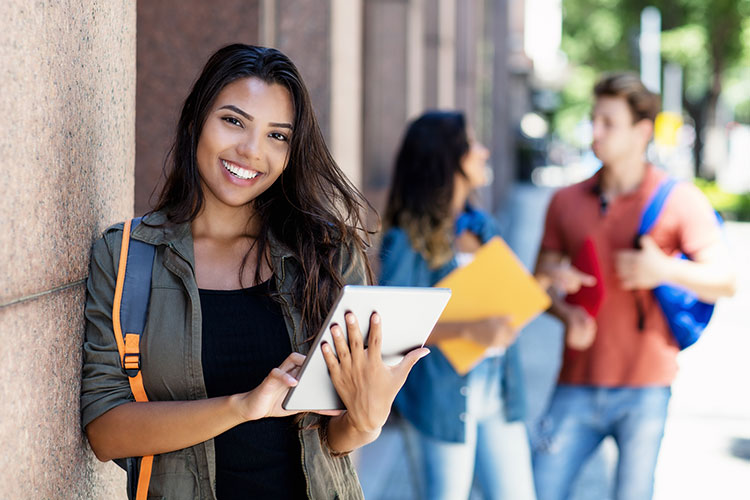 طالب مكسيكي يضحك مع كمبيوتر لوحي وأصدقاء في الخلفية أمام مبنى الجامعة